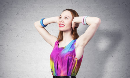Redhead beautiful girl closed her ears with hands. Joyful lady with pleasant smile in bright colorful dress and bracelets against grey wall background. Portrait of happy young woman looking up aside.の写真素材