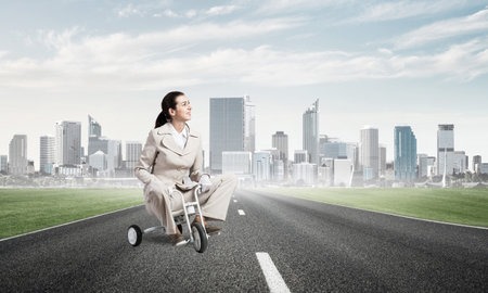 Beautiful young woman riding childrens bicycle on asphalt road. Businesslady in white business suit cycling small bike outdoor. Modern city on horizon. Professional career start concept.の写真素材