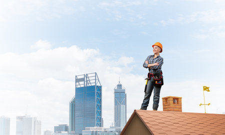 Attractive female construction worker in hardhat standing with folded arms. Confident young woman in checkered blue shirt standing on brick roof of building. Professional perspective and motivationの写真素材