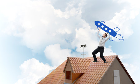 Conceptual image of young confident businessman in suit launching big blue rocket from his hand while standing on the top of brick roof with cloudy skyscape view on background.の写真素材