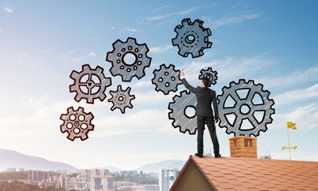 Young businessman standing on house roof and drawing gear mechanism. Mixed mediaの写真素材