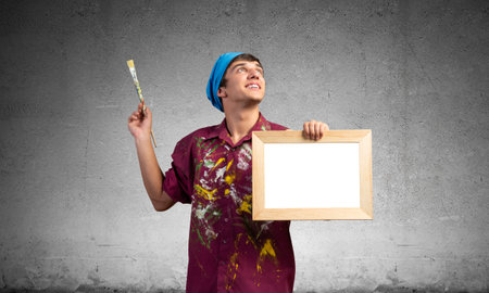 Young artist standing with blank white board in frame. Happy painter in dirty shirt and bandana with paintbrush on grey wall background. Creative hobby and artistic occupation concept with copy spaceの写真素材