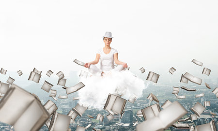 Woman in white clothing keeping eyes closed and looking concentrated while meditating on cloud among flying books with cityscape view on background.の写真素材