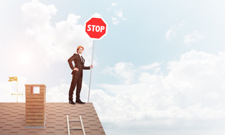 Engineer man standing on house roof and holding red prohibition sign. Mixed mediaの写真素材