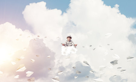 Young little boy keeping eyes closed and looking concentrated while meditating among flying paper planes in the air with cloudy skyscape on background.の写真素材
