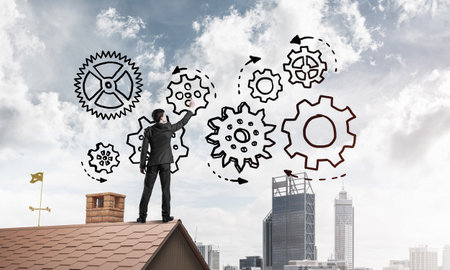 Young businessman standing on house roof and drawing gear mechanism. Mixed mediaの写真素材