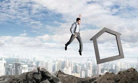 Jumping businessman crashing big house symbol with city view on background. 3D rendering.の写真素材