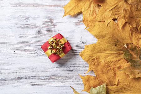Bright autumn composition with gift box and yellow maple leaves. Holiday present decorated golden ribbon bow lies on vintage wooden desk. Happy thanksgiving congratulation. Autumn sale advertising.の写真素材