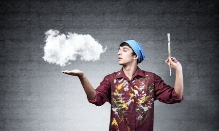 Young artist drawing realistic white cloud. Male painter in dirty shirt and bandana with paintbrush on grey wall background. Creative hobby and artistic occupation. Art classes concept with copy spaceの写真素材
