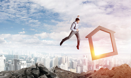 Jumping businessman crashing big house symbol with city view on background. 3D rendering.の写真素材