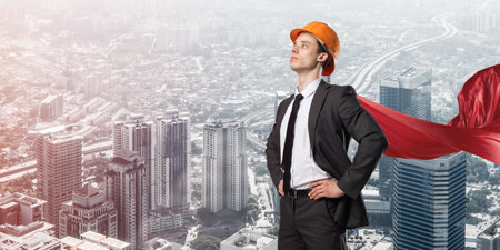 Confident engineer man in helmet and red cape against modern cityscape backgroundの写真素材