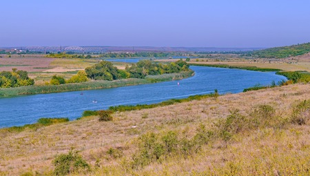 A wide river flowing between fields and low slopes covered with dry grass with peacefully grazing...