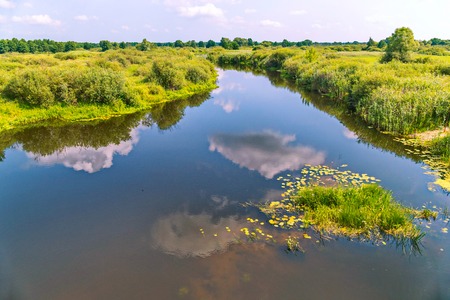 The clouds are reflected in the blue river, dividing the green grassy terrain into two shoresの写真素材