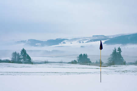 A golf course in Scotland on a snowy winter morning in December. View from a green with a blue flag, with trees, mist and mountains in the background.の写真素材