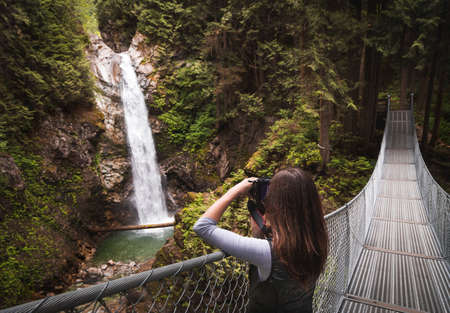 A young woman on a suspension bridge taking a photo of a waterfall.