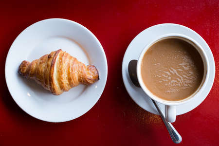 A croissant and a cup of coffee on a red table.の写真素材