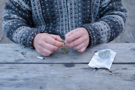 Close-up of a mans hands breaking up marijuana buds next to a dispensary bag and a joint.