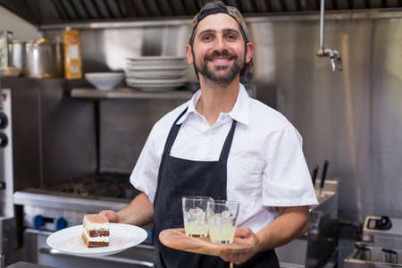 A happy smiling male chef holding tiramisu and limoncello in a restaurant kitchen.の写真素材