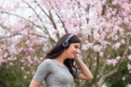 Young woman listening to music on wireless headphones in a park with cherry blossom trees.