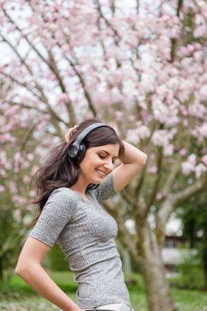Young woman listening to music on wireless headphones in a park with cherry blossom trees.の写真素材