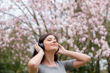 Young woman listening to music on wireless headphones in a park with cherry blossom trees.の写真素材