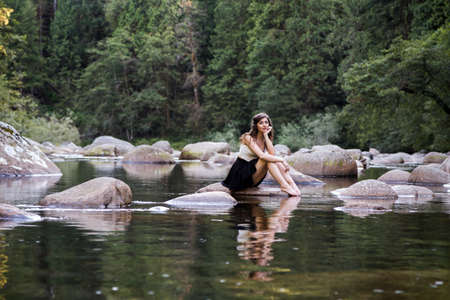 Young attractive brunette woman on a rock next to a pristine river in an evergreen forest.の写真素材