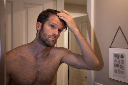 Handsome young caucasian man looking at himself in a bathroom mirror while examing his receding...