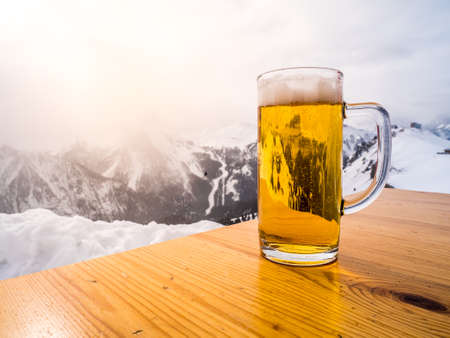 Glass or pint of a beer on a wood table on the top of the alps. Sun light is shining on the winter...