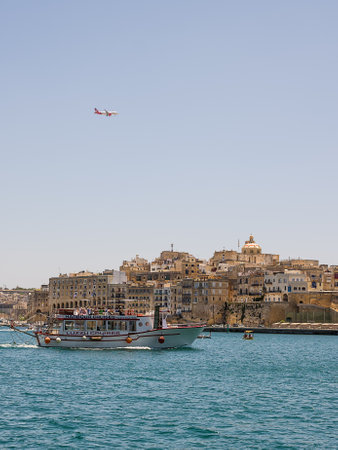 Valletta, Malta - 17 June 2023: Gulf between Valletta and Vittoriosa with passing boat with tourists and international plane in the skyのeditorial素材