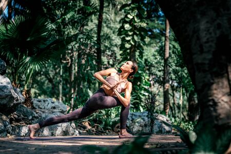 Full-length shot of caucasian woman standing in Crescent Lunge Twist pose, while practicing yoga outdoors, in a garden. Healthy lifestyle and relaxation conceptの写真素材