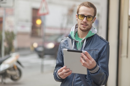 Young man with yellow glasses use iPad tablet computer on street, public space. Blurred backgroundのeditorial素材