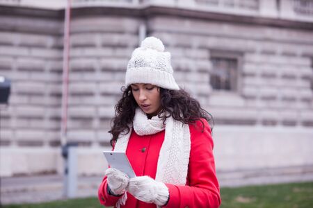 Smiling Woman in Red Coat and black hat and glasses, hold smartphone, Woman in red coat with smartphone in hands going through the city and looking shop windows. Urban Space, noise on imageの写真素材
