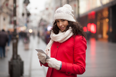 Beautiful Woman in Red Coat and and wool cap and gloves read messages on smartphone and smiling. Urban Space. Cold Weatherの写真素材