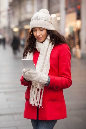 Beautiful Woman in Red Coat and and wool cap and gloves read messages on smartphone and smiling. Urban Space. Cold Weatherの写真素材