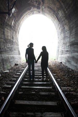 Couple walking hand in hand along the track through a railway tunnel towards the bright light at the other end, they appear as silhouettes against the lightの写真素材