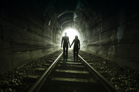 Couple walking hand in hand along the track through a railway tunnel towards the bright light at the other end, they appear as silhouettes against the lightの写真素材