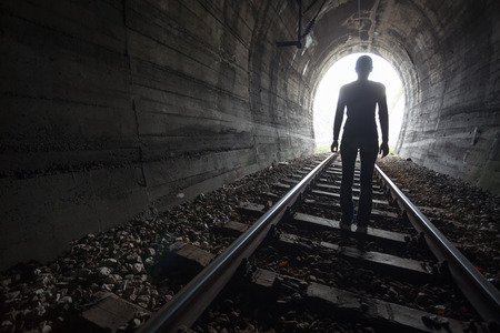 Man silhouetted in a tunnel standing in the center of the railway tracks looking towards the light at the end of the tunnel in a conceptual imageの写真素材