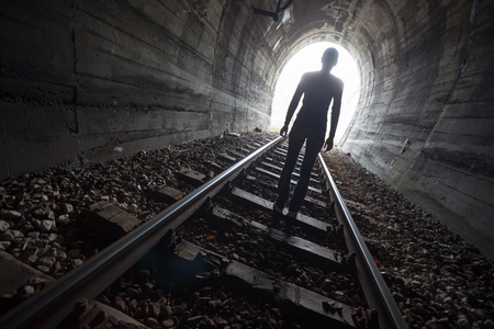 Man silhouetted in a tunnel standing in the center of the railway tracks looking towards the light at the end of the tunnel in a conceptual imageの写真素材