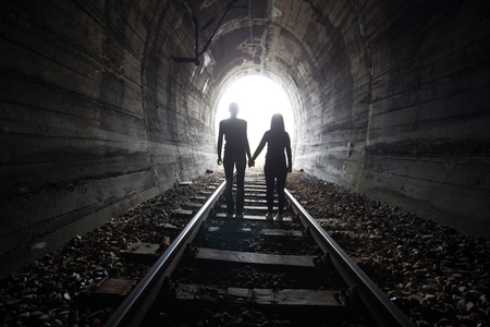 Couple walking hand in hand along the track through a railway tunnel towards the bright light at the other end, they appear as silhouettes against the lightの写真素材