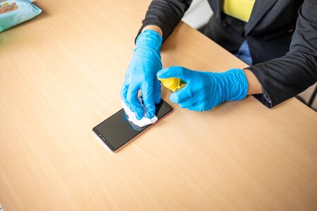 Woman's hand in blue gloves sanitizing cleaning smartphone mobile phone on wood table surface with wet wipesの写真素材
