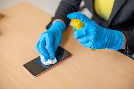 Woman's hand in blue gloves sanitizing cleaning smartphone mobile phone on wood table surface with wet wipesの写真素材