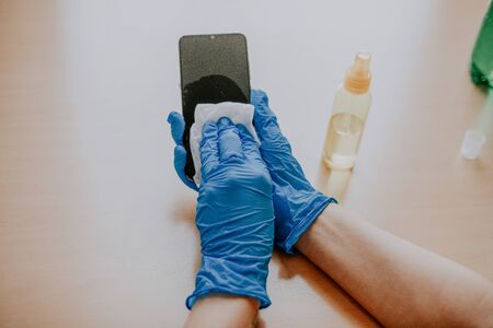 Woman's hand in blue gloves sanitizing cleaning smartphone mobile phone on wood table surface with wet wipes and alcoholの写真素材