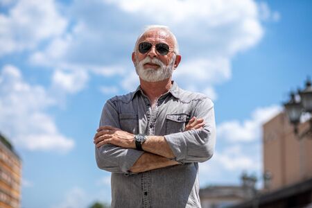 Old rich man with beard and sun glasse on street posing and smiling, portrait, blurred clouds backgroundの写真素材