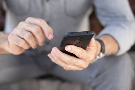 Old man in gray clothing seating on street and typing SMS on smartphoneの写真素材