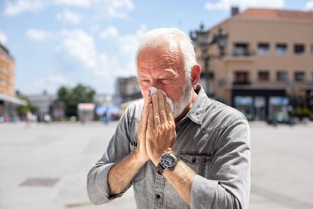 Old man coughs and sneezes into a handkerchief on street, outdoor, hot summer, allergies and illness conceptの写真素材