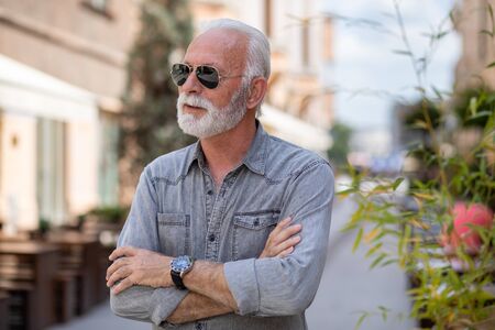 Happy rich old man with beard and sun glasses on street posing and smiling, portrait, blurred city street backgroundの写真素材
