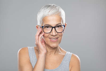 Beautiful smiling cute senior woman with short white hair and glasses posing in front of gray backgroundの写真素材