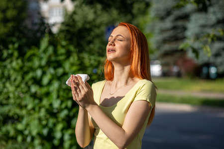 Young redhair woman sneezing in front of blooming tree. Pollen Allergy symptoms, green park in backgroundの写真素材