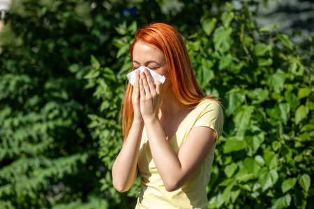Young redhair woman sneezing in front of blooming tree. Pollen Allergy symptoms, green park in backgroundの写真素材