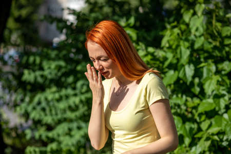 Young redhair woman sneezing in front of blooming tree. Pollen Allergy symptoms, green park in backgroundの写真素材